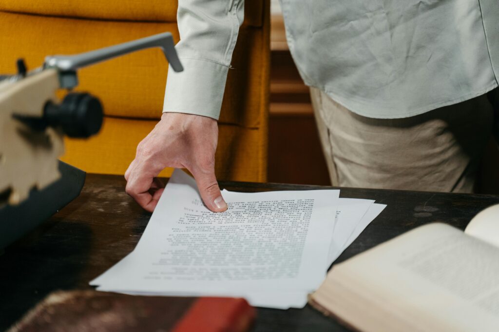 교수 논문 중복게재 징계처분 무효 승소사례 Close-up of a hand reaching for a document on a desk with a typewriter and book.